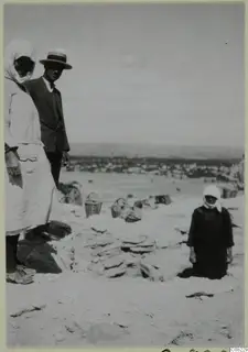 Woman and man in a straw hat beside stacked stones and wicker baskets on sandy ground, with a figure in a headscarf at right