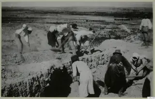 Workers digging a trench at an excavation, with a flat plain and low hills in the background.