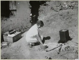 Woman crouching beside a round bowl on bare ground, apparently washing, outside a mud-walled building at Machairas