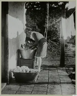 Woman washing cloth in a metal tub beside a whitewashed building wall, with laundry hanging on a line