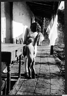 Woman leaning over a small naked child on a covered veranda beside a wooden table