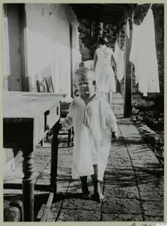Young child in a white nightshirt standing barefoot on a paved veranda beside a wooden table