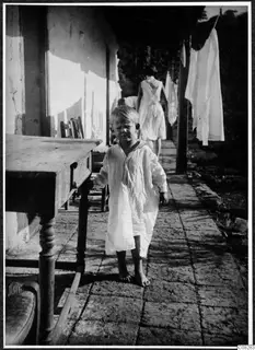 Young child in a loose white garment standing on a paved veranda beside a wooden table, with laundry hanging behind