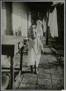 Young child in a white nightdress standing barefoot on a stone-paved veranda beside a wooden table