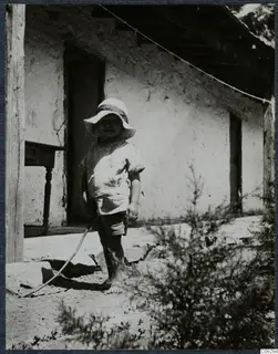 Young child in a wide-brimmed hat standing barefoot in front of a plastered building at Machairas