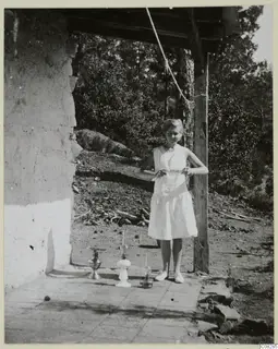 Young woman in a white dress stands beside three oil lamps on a paved terrace outside a building.