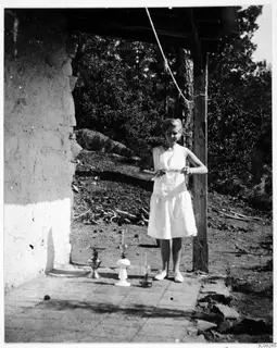 Woman in a light dress standing beside three oil lamps on a paved terrace next to a plastered mud wall.
