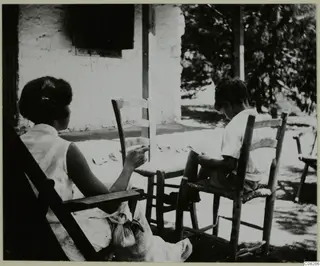 Two people sitting on wooden chairs on a veranda in front of a whitewashed building, one reading.