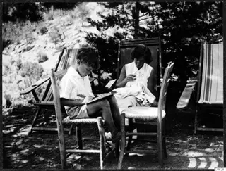 Two people sitting outdoors on wooden chairs: a boy writing in a book and a woman sewing white cloth, under pine trees.