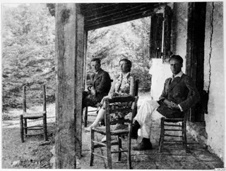 Three people seated on a shaded veranda of a whitewashed building at Machairas, with rush-seated chairs