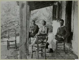 Three people seated on the veranda of a whitewashed building at Machairas, with rush-seated chairs around them.