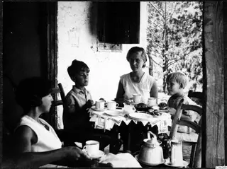 Four people seated at a table with cups, plates and food on a lace-edged cloth, on a veranda with trees beyond.