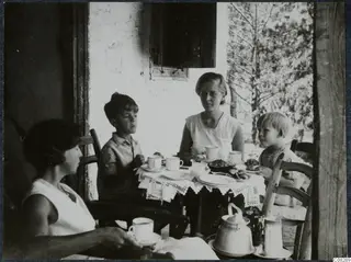 Four people seated at a table with cups, plates and a teapot on a shaded veranda beside a white wall