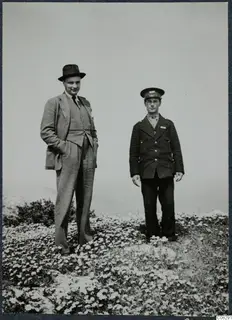 Two men standing in a field of small white flowers at Vouni, one in a suit and hat, one in a uniform cap.