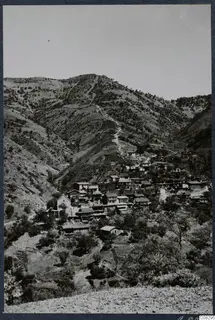 Village of houses clustered on a steep wooded mountainside, with a footpath climbing the slope above it.