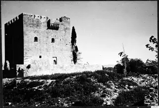 Square stone tower with battlements and small arched windows, with a tall cypress tree beside it, seen across a grassy slope.