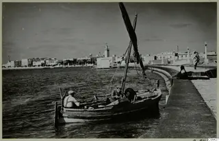Wooden boat with three men in calm water beside a curved stone quay, with a town of white buildings along the shore