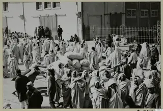 Crowd of people in pale hooded cloaks carrying sacks across a paved yard beside a large building