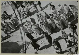Crowd of people in long pale hooded robes walking across open ground, with hand carts and sacks and ropes in the foreground