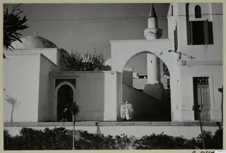 Whitewashed buildings with a dome, a tall tower and an arched passage, with two people in white robes walking past a hedge.