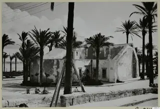 Whitewashed building with arched openings among palm trees, with people seated on a street corner, North Africa.