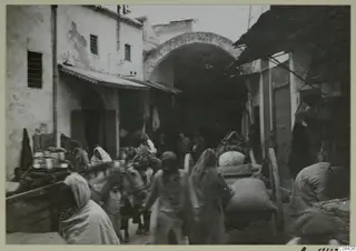 Street scene in North Africa with an arched passage, people, and a loaded donkey or mule.