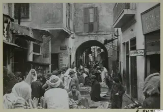 Crowded market street under an archway, with people and produce on low stalls, recorded as North Africa.
