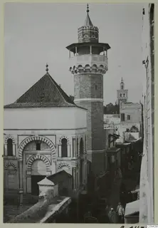 Mosque with a tall minaret and banded arches, with people in a narrow street, North Africa