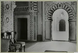 Interior of a building with a striped arch framing a studded wooden door, patterned wall panels and upholstered chairs in the foreground.