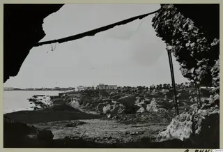 Rocky coastline and stones seen from under a dark rock overhang, with water and buildings on the horizon, North Africa.