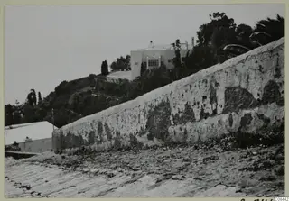 Weathered plaster wall running up a slope, with a white building and trees behind, North Africa