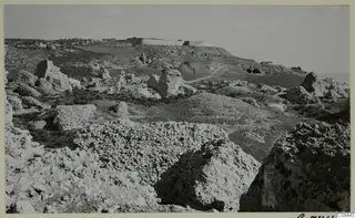 Rocky limestone ground with fallen stone blocks, a long walled structure on a hilltop and a small building, with the sea at right.