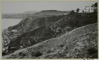 Rocky hillside above the coast with trees, a winding road and a white building on the upper right.