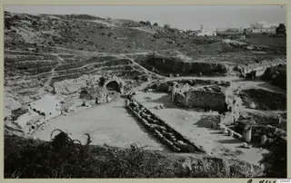 Ruined stone walls and a vaulted arch on a hillside site, with a white domed building in the distance