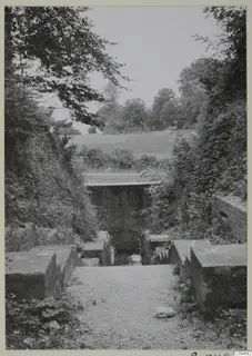 Stone-walled steps leading up a gravel path toward a brick-faced structure, with trees and bushes on both sides