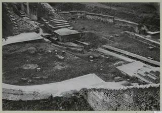 Excavated brick ruins with stepped brick structures, low walls and paved surfaces on a grassy site, seen from above