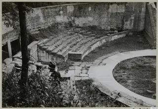 Curved brick-walled structure with tiered stone seating and a paved walkway, with two men in hats at the left edge