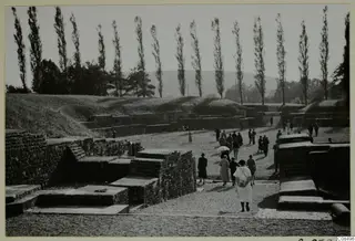 Visitors walking among stone ruins and low walls, with a grassy bank and tall poplars behind.