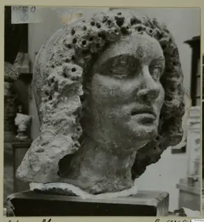 Sculpted stone head with curled hair pierced by many small holes, on a dark plinth