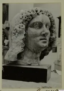 Female head in pale stone with curled hair pierced by drilled holes, on a dark wooden base