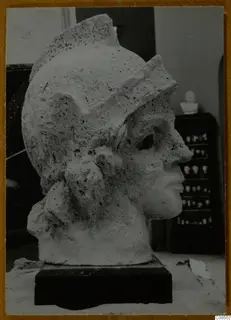Helmeted head sculpture in pale, pitted stone, shown in right profile on a dark rectangular base