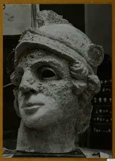 Carved stone head with a close-fitting cap, large hollow eye sockets and a damaged nose, on a dark base