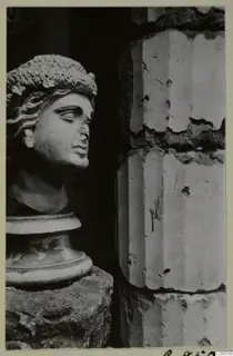 Carved stone head with wavy hair on a moulded base, set on a rough stone block beside a masonry wall