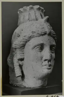 Carved stone head of a woman with wavy hair and a tall crown-like headdress, on a dark wooden stand.