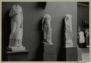 Three headless stone statues on pedestals against a dark wall, with more sculptures in a room to the right.