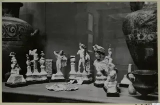 Small white figurines and a dark bowl displayed on a shelf, with large vessels behind them