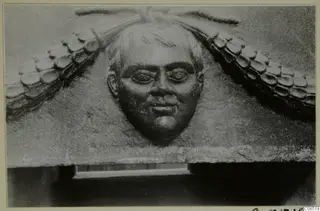 Carved stone relief of a man's face with curled hair, set between scale-patterned ornament on a block.
