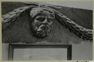 Carved bearded male face in relief, with feather-like shapes on either side, set into a wall above a framed opening