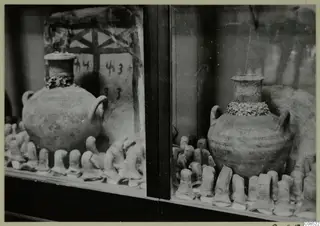 Two round jars with short necks and handles standing in a glass display case among rows of small clay objects