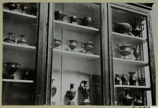 Dark vessels with handles and jugs displayed on shelves in a glass-fronted wooden cabinet.